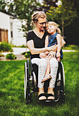 Eine querschnittsgelähmte Mutter hält ihr kleines Mädchen auf dem Schoß, während sie an einem warmen Sommernachmittag in ihrem Vorgarten im Rollstuhl sitzt: Edmonton, Alberta, Kanada.