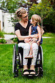 A paraplegic mom holding her little girl in her lap while sitting in her wheelchair in her front yard on a warm summer afternoon: Edmonton, Alberta, Canada.