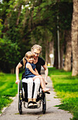 Eine querschnittsgelähmte Mutter hält ihr kleines Mädchen auf dem Schoß, während sie es an einem warmen Sommernachmittag in ihrem Rollstuhl die Straße hinunterfährt: Edmonton, Alberta, Kanada.