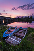 Two old wooden boats on the banks of the Shannon River at sunset with a stone bridge in the background; Montpellier, County Limerick, Ireland