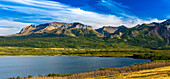 Panorama of mountain range along a lakeshore with blue sky and clouds, Waterton Lakes National Park; Waterton, Alberta, Canada