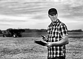 Ein Landwirt nutzt sein Tablet, um bei der Weizenernte zu helfen, während im Hintergrund ein Getreidewagen arbeitet: Alcomdale, Alberta, Kanada
