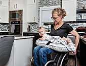 A paraplegic mother holding her baby on her lap, in her kitchen, while pushing in her wheel chair: Edmonton, Alberta, Canada