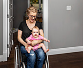 Eine querschnittsgelähmte Mutter, die ihr Baby auf dem Schoß hält, während sie sich im Rollstuhl durch ihr Haus bewegt:: Edmonton, Alberta, Kanada