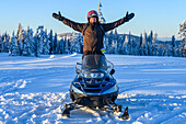 Man stands on his snowmobile with arms outstretched while looking at the camera on the pristine slopes of the Rocky Mountains; Sun Peaks, British Columbia, Canada