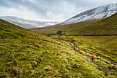 Lone female hiker in red raincoat carrying a red backpack walking along a valley trail leading to snow-covered mountains on a cloudy winter day, Galty Mountains; County Tipperary, Ireland
