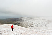 Wanderin in roter Jacke beim Wandern auf einem schneebedeckten Berg im Winter bei schlechtem Wetter, Galty Mountains; County Tipperary, Irland