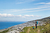Woman and dog hiking on trail in the Burren looking out at the sea on a sunny summer day; Fanore, County Clare, Ireland
