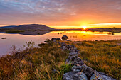Kleine Steinmauer, die zu einem See mit einem Berg und einem Sonnenaufgang führt, der sich im Wasser spiegelt, Burren National Park; County Clare, Irland
