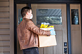 Ein junger Mann steht mit einer Kiste voller Lebensmittel vor der Tür eines Hauses während der Covid-19-Weltpandemie; Edmonton, Alberta, Kanada