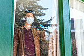 Ein junger Mann trägt eine Maske und steht während der Covid-19-Weltpandemie hinter einem Fenster in seinem Haus; Edmonton, Alberta, Kanada