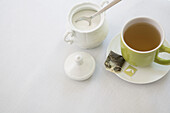 Gebrauchter Teebeutel auf Untertasse mit Tasse Tee in grüner Tasse mit Zuckerdose und Löffel, Studioaufnahme