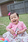 Porträt eines kleinen Mädchens in Rosa, das mit einem Osterkorb im Garten auf dem Gras sitzt und lacht