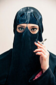 Close-up portrait of young woman wearing black, muslim hijab and muslim dress, holding cigarette and smoking, looking at camera, eyes showing eye makeup, studio shot on white background