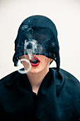 Close-up portrait of young woman wearing black, muslim dress and black, hijab covering part of head, while blowing smoke rings from red lips, studio shot on white background