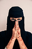Close-up portrait of woman wearing black muslim hijab and muslim dress looking at camera, with hands praying and showing arms and hands painted with henna in arabic style, studio shot on whtie background