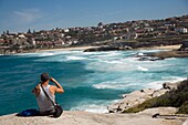Mann auf einem Felsen sitzend mit Blick auf Mackensies und Tamarama Bay, in der Nähe von Bondi Beach