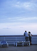 Couple Looking Out From Deck Of Cruise Ship At Dusk