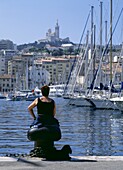 Lady Sitting Beside The Harbor With Her Dog With Notre Dame De La Garde Cathedral On The Hill Behind
