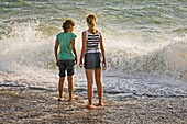 Zwei junge Mädchen stehen am Strand und halten ihre Füße ins Wasser.