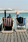 Zwei Personen entspannen sich auf Liegestühlen am Brighton Pier