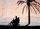 Silhouette Of Father And Two Sons On Moped Going Past Shadow Of Date Palm On City Walls