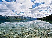 Snowcapped Mountains Reflected In Lake Wakatipu With Clear Water And Pebbles