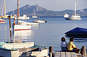 Two Young Children Sitting On A Dock In Port De Mollenca