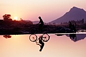 Fahrradfahrer beim Überqueren eines seichten Flusses bei Sonnenuntergang mit Frauen im Hintergrund, die Wäsche waschen