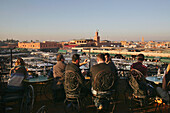 Touristen beim Teetrinken auf dem Balkon eines Cafés mit Blick auf den Nachtmarkt Djemaa El Fna.