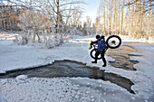 Fahrradfahrer überquert das offene Wasser des Campell Creek, während er ein Snow Bike im Far North Bicentennial Park in der Nähe von Anchorage, Süd-Zentral-Alaska, Winter, trägt