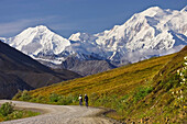 Two Women Ride Bikes Over Thorofare Pass With Mt. Mckinley In The Background In Denali National Park, Alaska