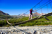 A Woman Walks On A Suspension Bridge Across College Creek To Gulkana Glacier, Southcentral Alaska, Summer/N