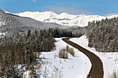 A Windy Hilly Road In The Winter With Snow Covered Hills, Trees And Mountains With Blue Sky; Alberta, Canada