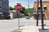 A stop sign and crosswalk in a small town, and view down a street of shops and parked cars. 