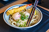 A dish of noodles, vegetables and fish and a tempura prawn, chopsticks. 