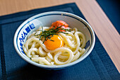 A dish of noodles, vegetables and fish with a yellow egg yolk. 