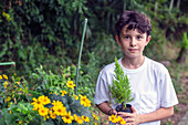 A boy holding a small tree sapling in a pot, standing in a garden. 