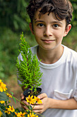 Ein Junge hält einen kleinen Bäumchen in einem Topf, stehend in einem Garten. 