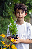 Ein Junge, der einen kleinen Baum in einem Topf hält, steht in einem Garten. 