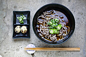 A black china bowl of noodles and broth, sliced green chilli and a dish with quail's eggs and sliced vegetables.