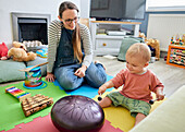 Kleinkind spielt mit Musikinstrument im Spielzimmer, Mutter sitzt in der Nähe