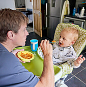 Father feeding toddler in high chair in kitchen