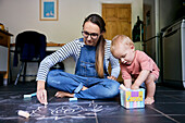 Mutter und Kleinkind malen gemeinsam mit Kreide auf einem gekachelten Küchenboden