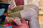 Close up of grandfather reading to toddler, view of the child's bare feet and grandpa's knees. 