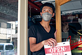 Ein Mann mit einer Gesichtsmaske hängt ein Schild mit der Aufschrift Open an die Tür eines italienischen Restaurants. Zweisprachig, Italienisch und Englisch. 