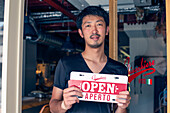 Ein Mann hängt ein Schild "Open" an eine Restauranttür. Zweisprachig, italienisch und englisch. 