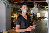 A man working in a restaurant, wearing a face mask, by an open kitchen, holding a digital tablet. 