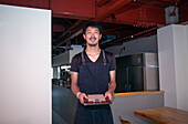 A man in a restaurant wearing an apron, holding a clipboard and menu, smiling in greeting. 