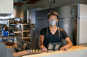 Ein Mann mit einer Gesichtsmaske an der Theke einer Restaurantküche, der ein digitales Tablet benutzt, der Besitzer oder Manager. 